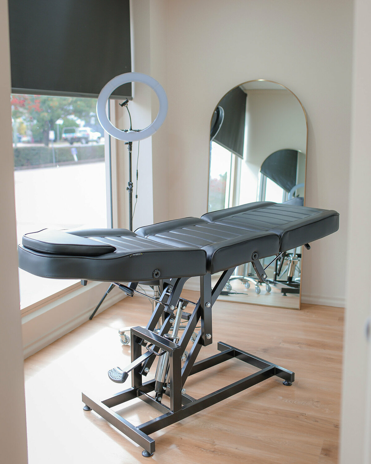 An adjustable studio chair/table sits in the middle of a room. Behind is a floor mirror and ring light.