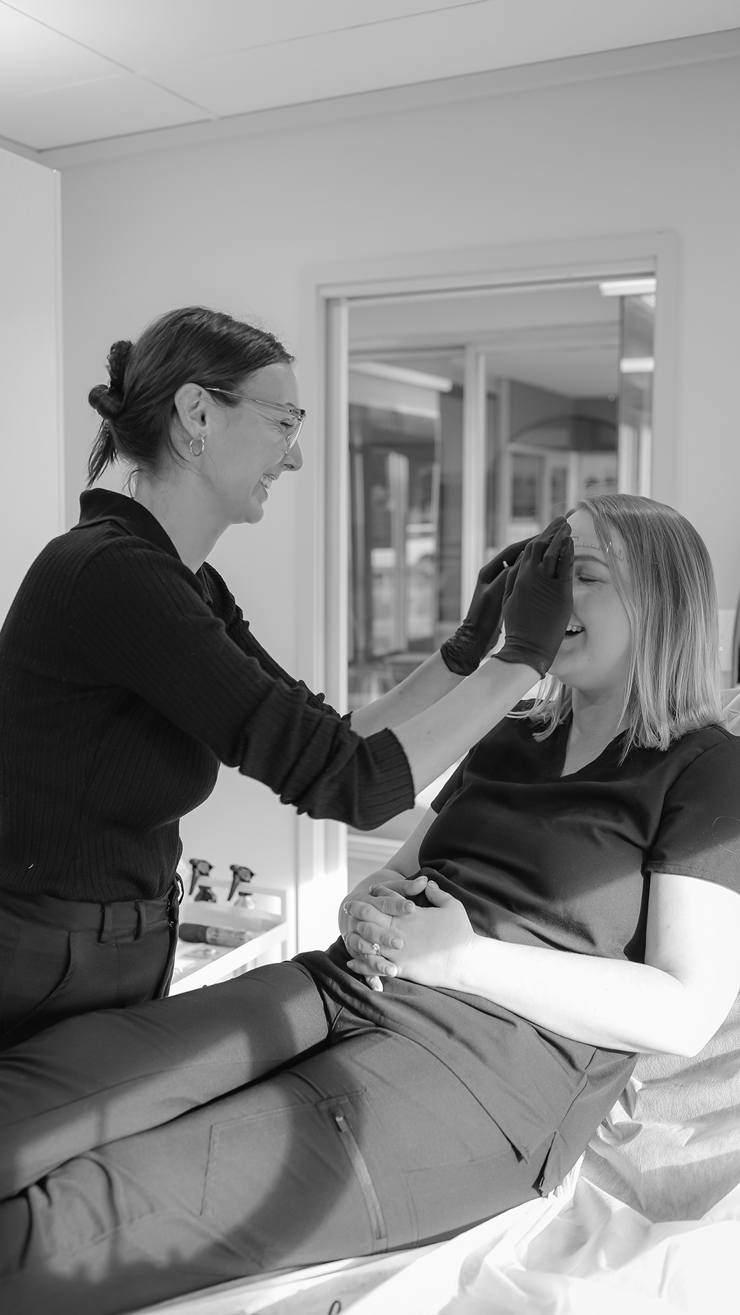 Lena is placing an eyebrow stencil on a client. They are both happy and laughing.